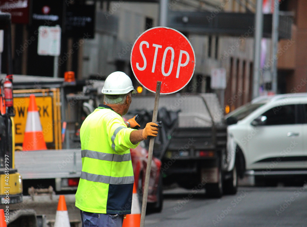 Male construction road worker holding a stop sign and directing traffic on the street. Traffic management - obrazy, fototapety, plakaty 