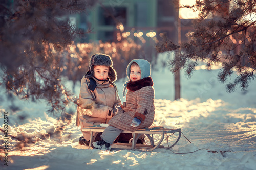 Cute little boy in earflaps hat and his baby sister in blue bonnet and retro coat are sitting on the old sledge in the winter park in Russia during the sunset. Image with selective focus and toning