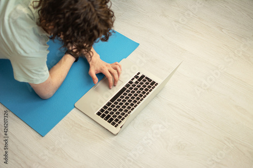 The concept of a group of employees meeting by video call. Side view. A Caucasian, curly-haired adult man is propped on his elbows at a remote work meeting, on a rubber yoga mat on a wooden floor.