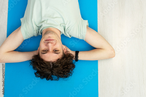 The concept of a happy man who engaged in psychological self-development. Caucasian adult, curly-haired man looking at the camera on a yoga mat on a wooden floor, an electronic bracelet on his hand.