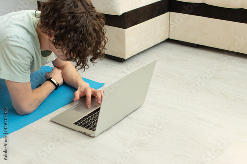 The concept of remote work during quarantine in a comfortable position.Adult Caucasian curly, with stubble man works on his own laptop while lying on a rubber yoga mat, which is on the wooden floor.