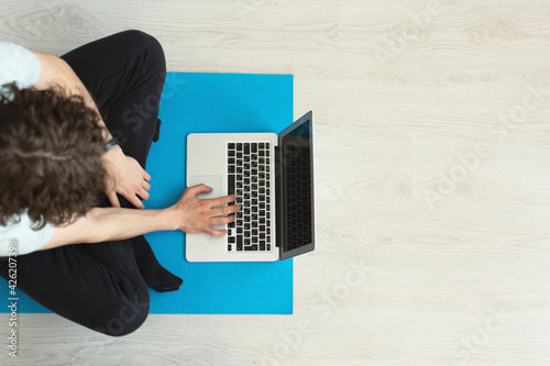 The concept of working from home during the quarantine period. Adult Caucasian curly-haired man sitting cross-legged on a rubber yoga mat on floor doing remote work on the computer. View from above.