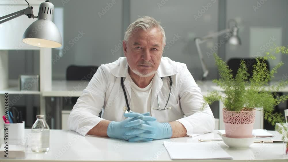An elderly professor of medical sciences sits at his desk, his gloved hands resting on the table An elderly professor of medical sciences sits at his desk, his gloved hands resting on the table