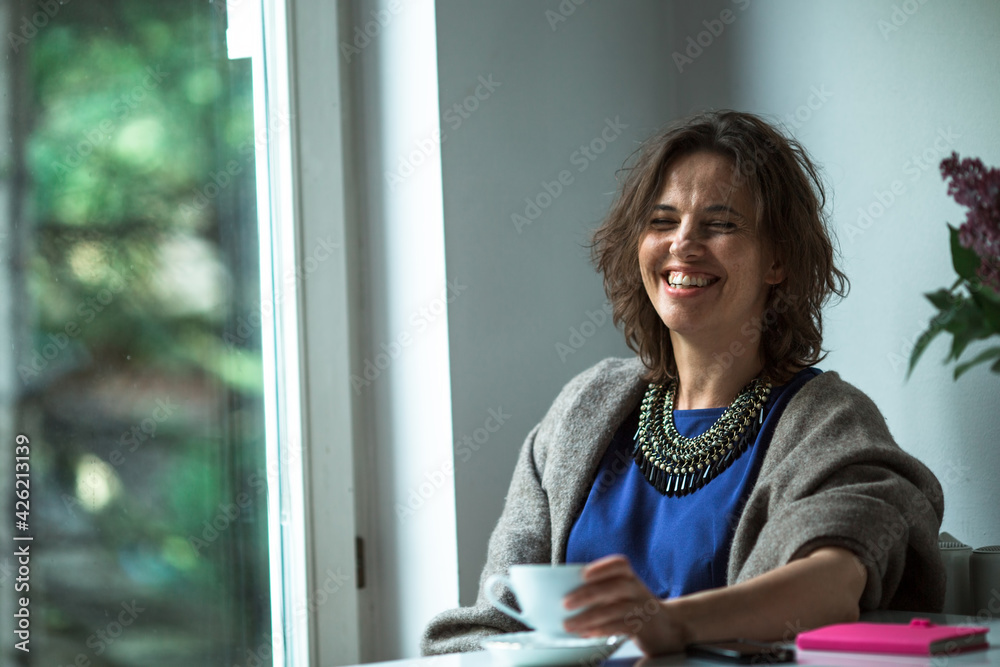 A woman with cup of tea sitting at a table near the window in her home. A woman with cup of tea sitting at a table near the window in her home.