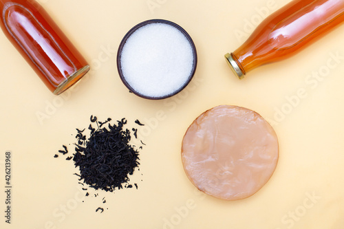 Two bottles of kombucha tea, scoby, brew and bowl of sugar on yellow pastel background. Ingredients for preparing healthy fermented drink. Flatlay mockup photo