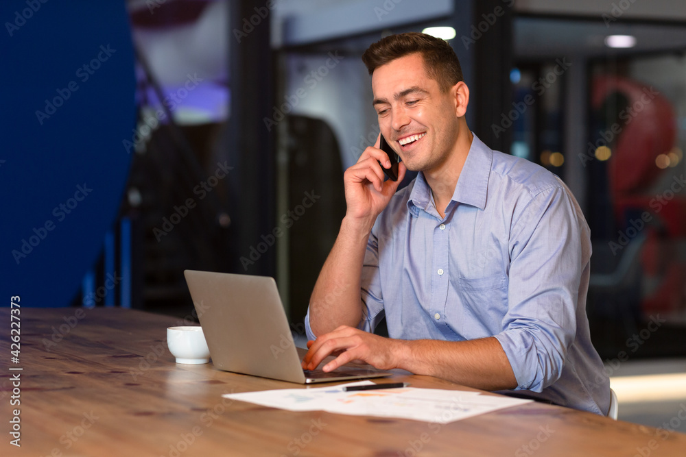 Portrait of happy casual caucasian businessman talking on smartphone at desk using laptop