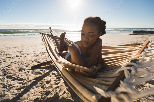 Happy african american woman lying in hammock on beach using tablet photo