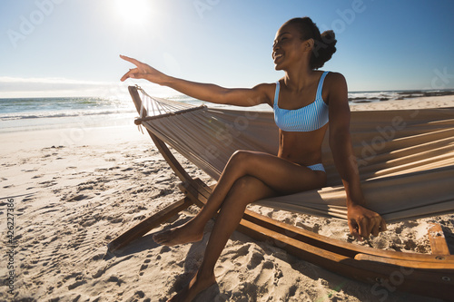 Happy african american woman sitting in hammock on beach pointing