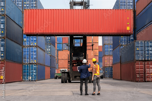 Inspector engineer in hard hat with Female dock worker standing in front of Reach stacker vehicle and used laptop checking cargo freight container to command transportation in logistic shipping yard