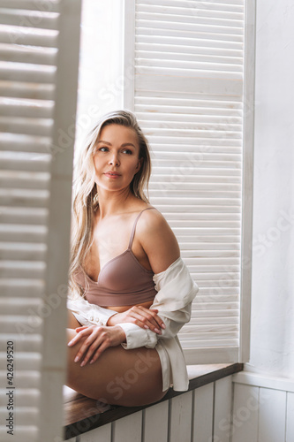 Blonde sensual young woman 35 year plus with long hair in underwear and white shirt sitting on window sill at the bright interior
