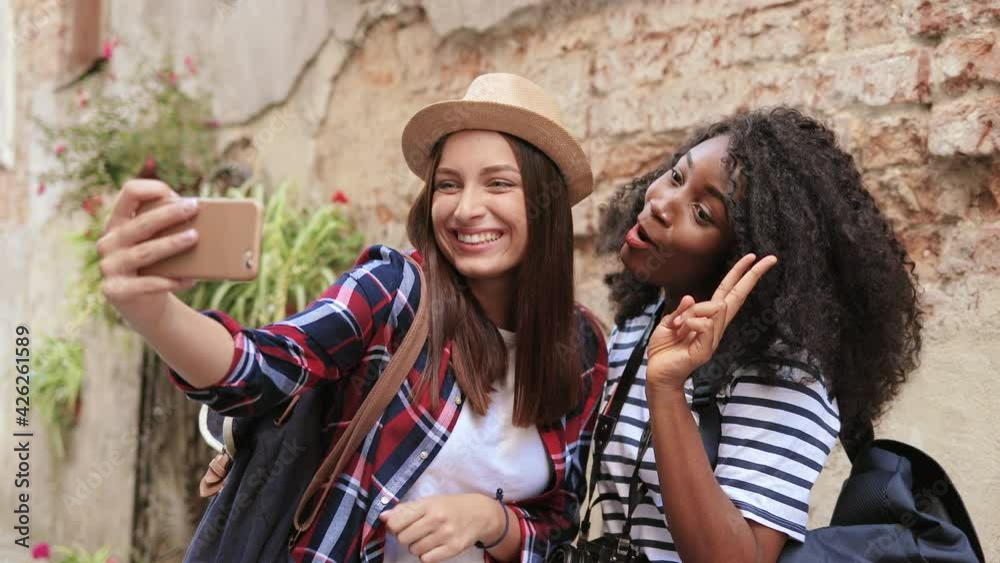 Young beautiful Caucasian girl in hat makes joint photo on smartphone with her African-American girlfriend with curly hair. Young beautiful Caucasian girl in hat makes joint photo on smartphone with her African-American girlfriend with curly hair.