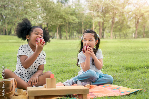 Happy cheerful ethnic girls eat apple together at outdoors park , Relationship little kids, Diverse ethnic concept.