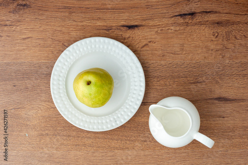 Top view of green pear on a white ceramic plate and empty milk jug on wooden table.