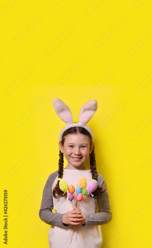 Funny happy girl with Easter bunny ears is holding colorful Easter eggs on a yellow background. Happy Easter! Copy space.Vertical photography