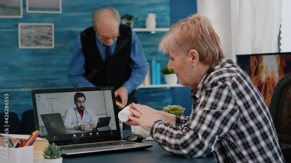 Old woman sitting at table at home looking at laptop answering question about health talks with therapist by videoconference call application, holding pills flask Remote medical consultation concept Old woman sitting at table at home looking at laptop answering question about health talks with therapist by videoconference call application, holding pills flask Remote medical consultation concept