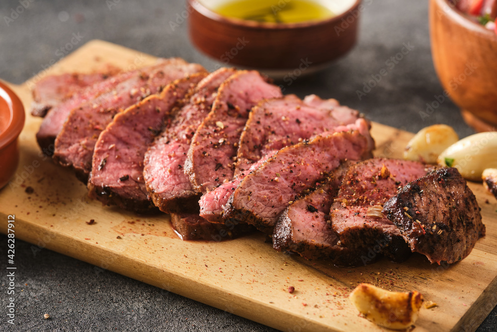 Sliced BBQ steak medium rare with salad and garlic on the grey background. Hip steak on wooden board Sliced BBQ steak medium rare with salad and garlic on the grey background. Hip steak on wooden board