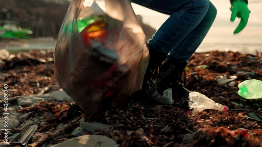 A volunteer walks along a dirty beach, in rubber gloves, picks up garbage and puts it in a plastic bag. Slow motion. Soft focus. Close up of feet.