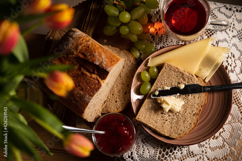 Frühstück ist fertig! Brot ist geschnitten, mit Butter geschmiert, Glas Tee , Weintrauben, Tulpen und Erdbeere Marmelade  stehen daneben auf dem Holztisch  Mit weißen Tischdecke dekoriert. Rustic photo