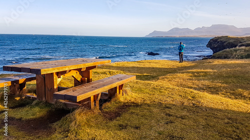 A resting bench situated at the shore of a steep cliff, overgrown with grass. In the back a young man is standing, and looking at the endless sea. Some taller mountains in the back.