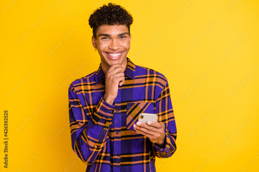 Photo of young afro guy happy positive smile hand touch chin think browse cellphone isolated over yellow color background Photo of young afro guy happy positive smile hand touch chin think browse cellphone isolated over yellow color background