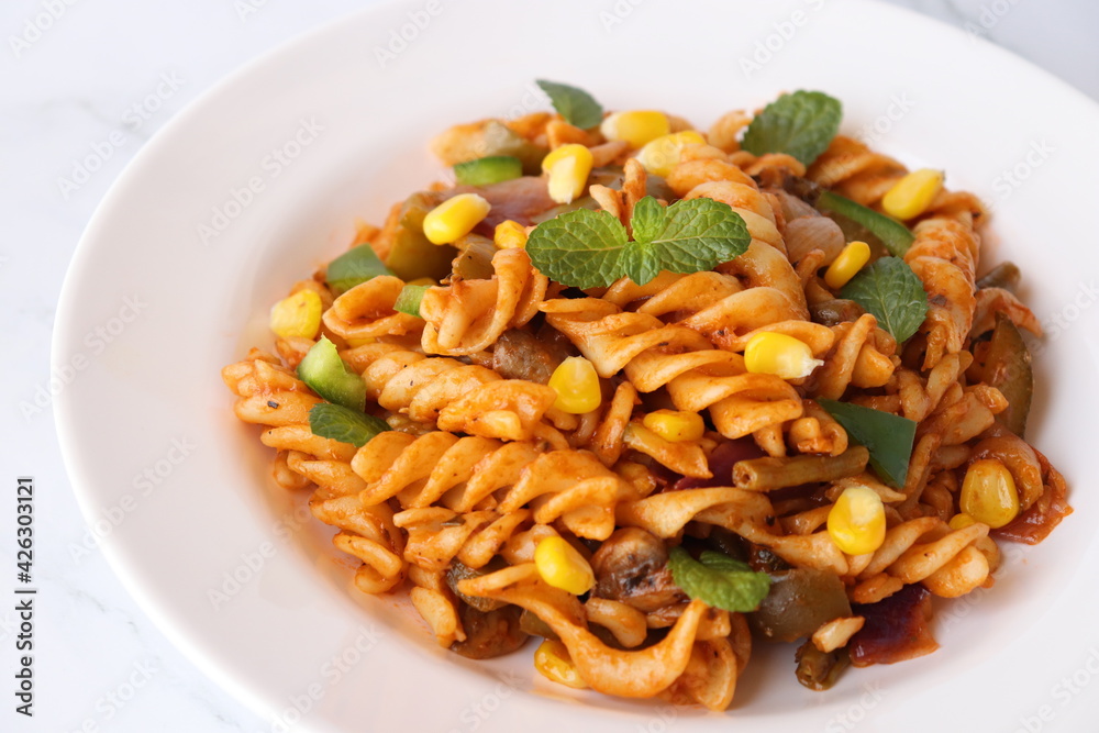 Whole wheat fusilli Pasta Arrabbiata with vegetables. Vegetable Pasta with zucchini, mushrooms, corn, and peppers on a white plate. on a white marble table with cutlery. copy space.