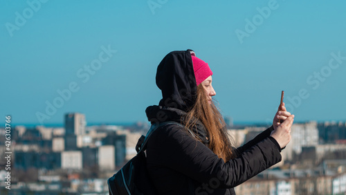 Young confident woman in pink hat with smartphone in hand calling friends walking on city background blue sky. Stylish millennial girl outdoor lifestyle authentic photo. Real people, solo travelling