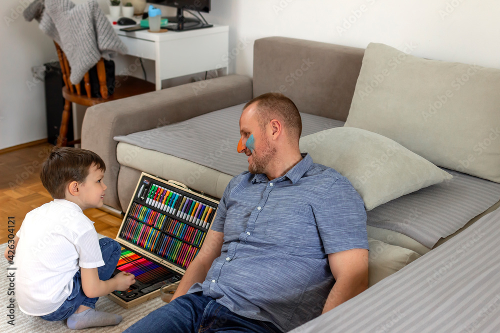 Obraz premium Cute little boy making colorful face on his father. Photo of a young father and his son enjoy the painting at home. Shot of a father and son doing arts and crafts at home. Dad and son having fun.