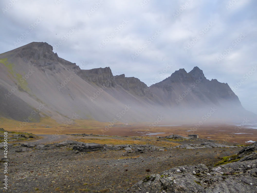Tall mountains emerging from the sea. The mist stays low, covering the lower parts of the mountains. Steep and harsh slopes. The ground is covered with rocks and pebbles with some grass in between.