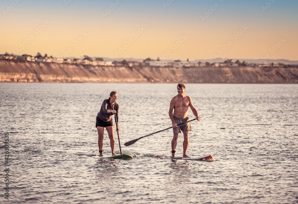 Naklejka premium Mature couple on SUP, stand up paddle board, having fun on quiet sea at sunset