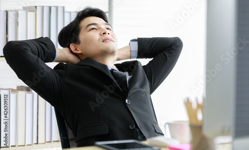 Asian young handsome black short hair male successful businessman entrepreneur wears formal suit with necktie and gray shirt sit smile relax stretching hold hands on head rest on company office chair