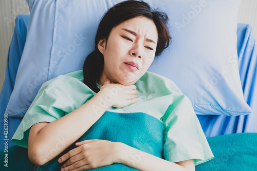 Asian young long hair sick ill girl patient wears green hospital uniform lay down on bed covered by blanket hold hand on chest sudden suffering from cough and pain in private ward room