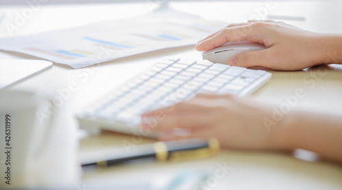 Closeup shot of female hand holding white wireless mouse while working with computer keyboard and chart graph document on office table with blurred foreground and background