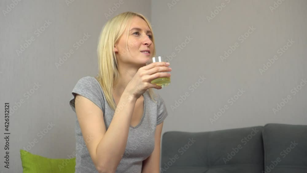 Young beautiful blonde woman drinking lemonade or juice at home. A woman looks away while sitting in an apartment. Young beautiful blonde woman drinking lemonade or juice at home. A woman looks away while sitting in an apartment.