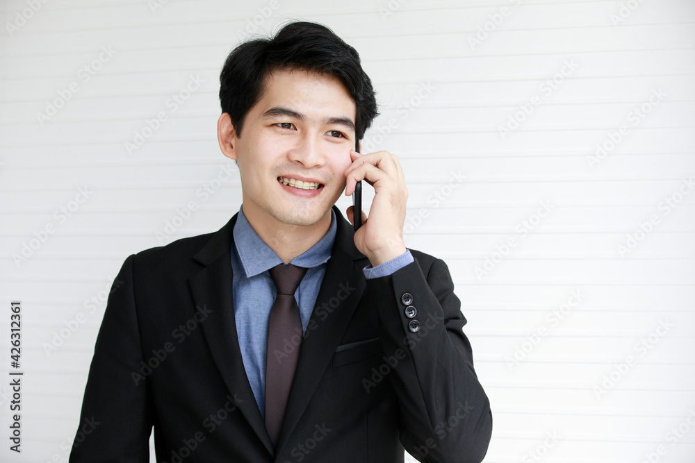 Portrait shot of Asian young successful male ceo businessman wears black formal suit stand smiling talking hold smartphone in hand has conversation with client and customer in front white wall Portrait shot of Asian young successful male ceo businessman wears black formal suit stand smiling talking hold smartphone in hand has conversation with client and customer in front white wall