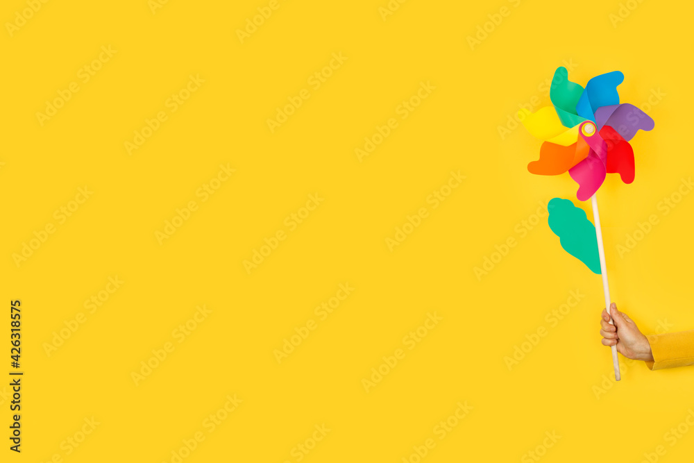 Fototapeta premium Mano de mujer sosteniendo un molinete molino de viento de colores sobre un fondo amarillo liso y aislado. Vista de frente. Copy space