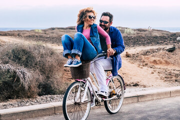 Young playful adult couple man and woman have fun together in outdoor leisure activity riding a single bike and laughing a lot - active people enjoy