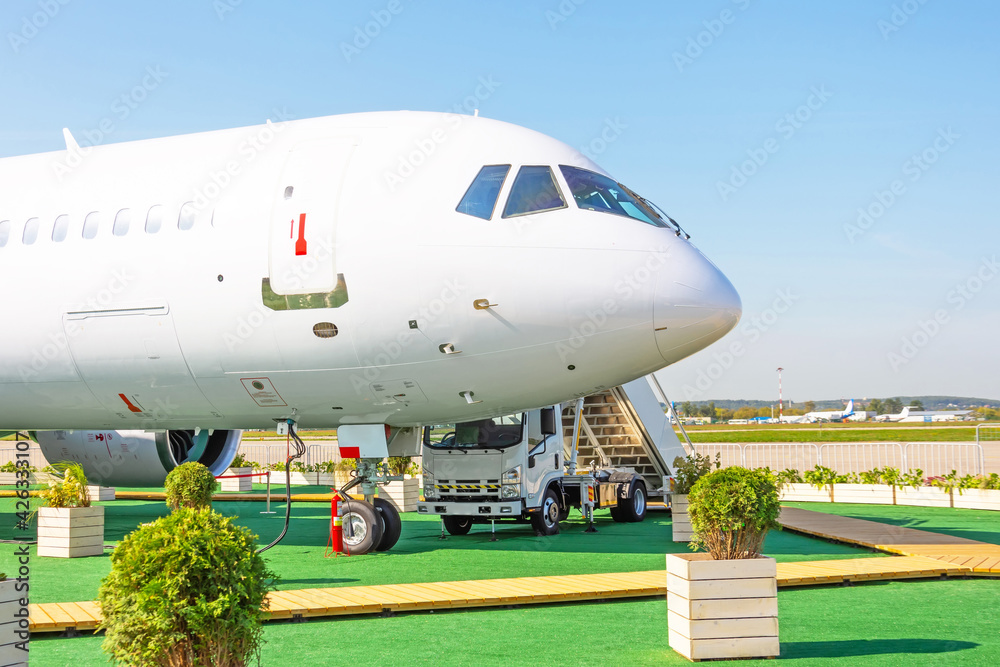 Airplane is parked among the trees and bushes of greenery with a walkway for passenger boarding, a view of the nose and cockpit.