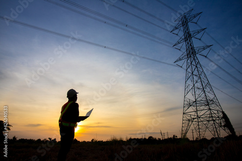 Silhouette of electrical engineer or technician standing and watching at the electric power station to view the planning work by producing electricity at high voltage electricity poles at the sunset.