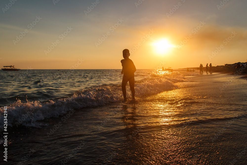 Silhouette of boy running barefoot along the shoreline on the beach at sunset in the summer