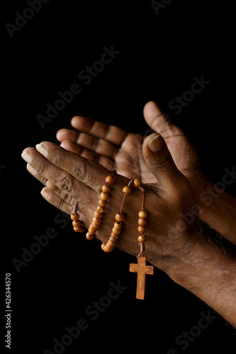 Praying hand of an old Indian Catholic man with wooden rosary isolated on a plain black background. (ID: 426344570)