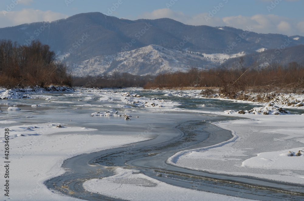 A frozen river in a wintry landscape. Winter landscape with forest, cloudy sky and sun.