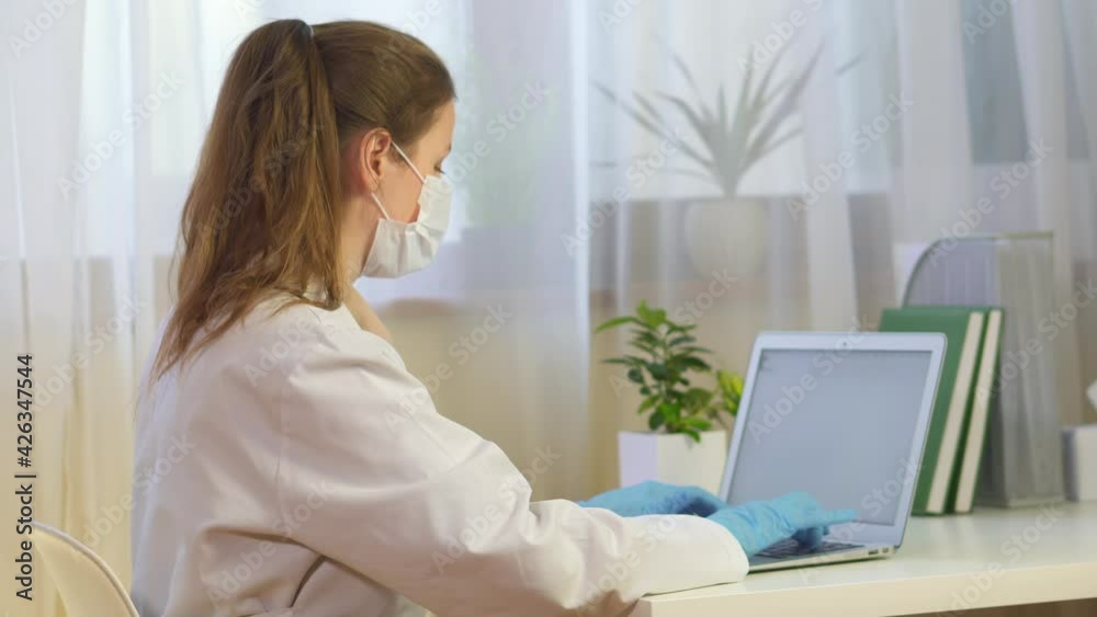 nurse works with a laptop in a mask and gloves. The doctor was distracted, looking into the frame. The doctor works online. Telemedicine doctor responds to a message online and looks into the frame. nurse works with a laptop in a mask and gloves. The doctor was distracted, looking into the frame. The doctor works online. Telemedicine doctor responds to a message online and looks into the frame.