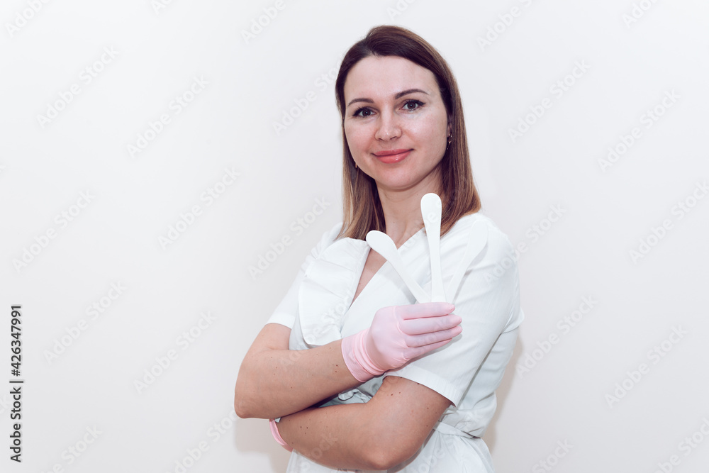 Doctor beautician in a white medical gown on a gray background holds a cosmetic spatula with place for text or logo. A woman in a white medical gown holds a white plastic cosmetic spatula. Doctor beautician in a white medical gown on a gray background holds a cosmetic spatula with place for text or logo. A woman in a white medical gown holds a white plastic cosmetic spatula.