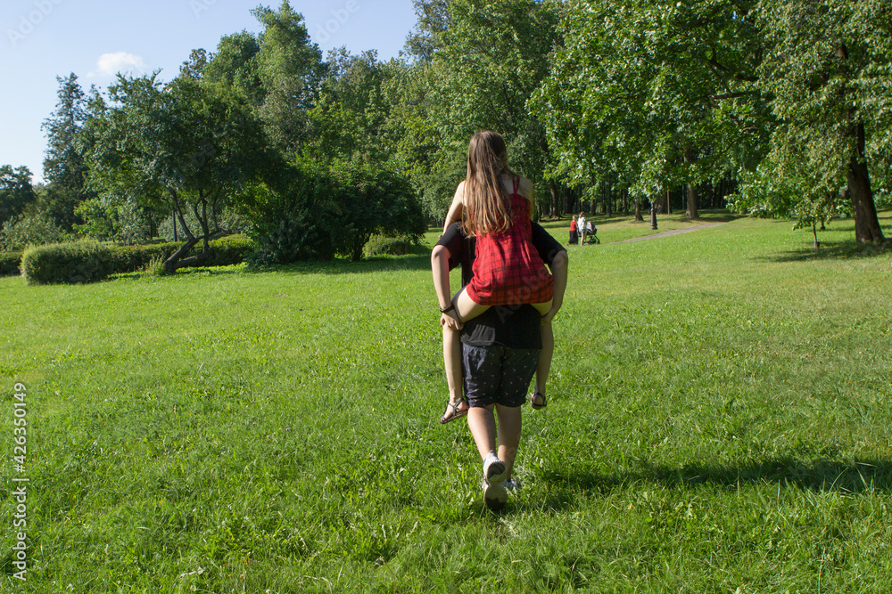 A young boy teenager is carrying his girlfriend on his back in a summer park. A loving couple frolics and has fun in nature. A young boy teenager is carrying his girlfriend on his back in a summer park. A loving couple frolics and has fun in nature.