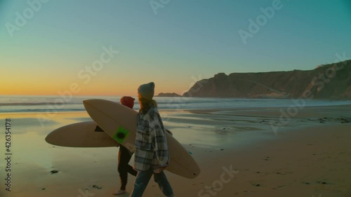 Incredible inspiring wide shot of two female friends walk barefoot in casual clothes hold surfboards on beautiful epic sunset beach. Romantic travel wanderlust concept. Millennial surfing adventure 
