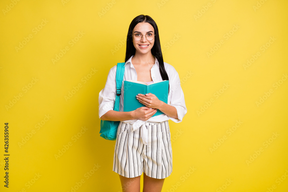 Photo of happy positive attractive woman hold book wear backpack isolated on shine yellow color background Photo of happy positive attractive woman hold book wear backpack isolated on shine yellow color background