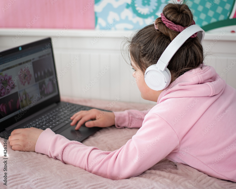 Dziewczynka pracuje zdalnie w domu przed komputerem ze słuchawkami na uszach leżąc na łóżku. - obrazy, fototapety, plakaty 