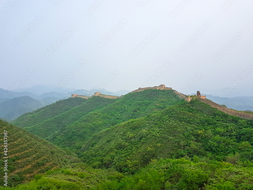 A panoramic view on an unrenewed Gubeikou part of Great Wall of China. The wall is spreading on tops of mountains. Many watchtowers on the peaks. Dense forest around it. World wonder. Tradition