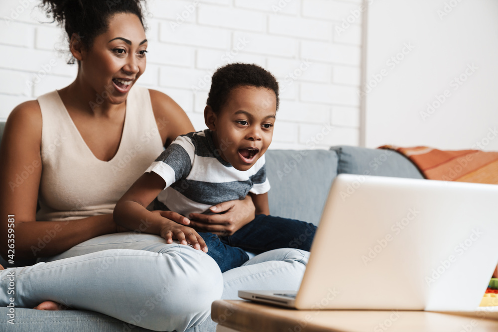 Fototapeta premium Black excited mother and son using laptop while sitting on sofa at home