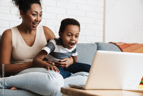 Black excited mother and son using laptop while sitting on sofa at home